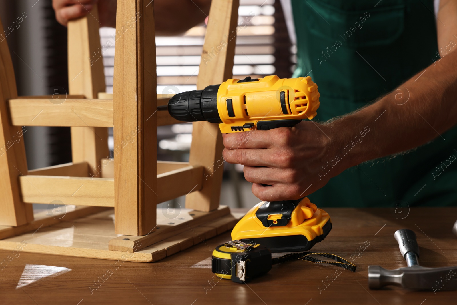 Photo of Man repairing wooden stool with electric screwdriver indoors, closeup