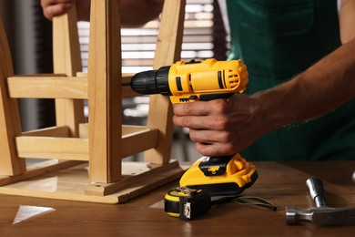 Photo of Man repairing wooden stool with electric screwdriver indoors, closeup