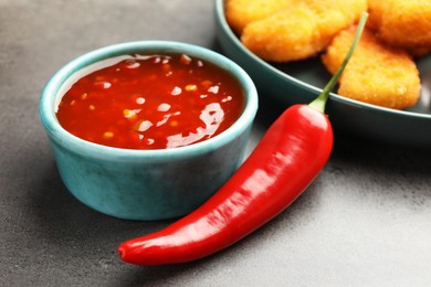 Photo of Hot chili sauce in bowl and pepper on grey textured table, closeup