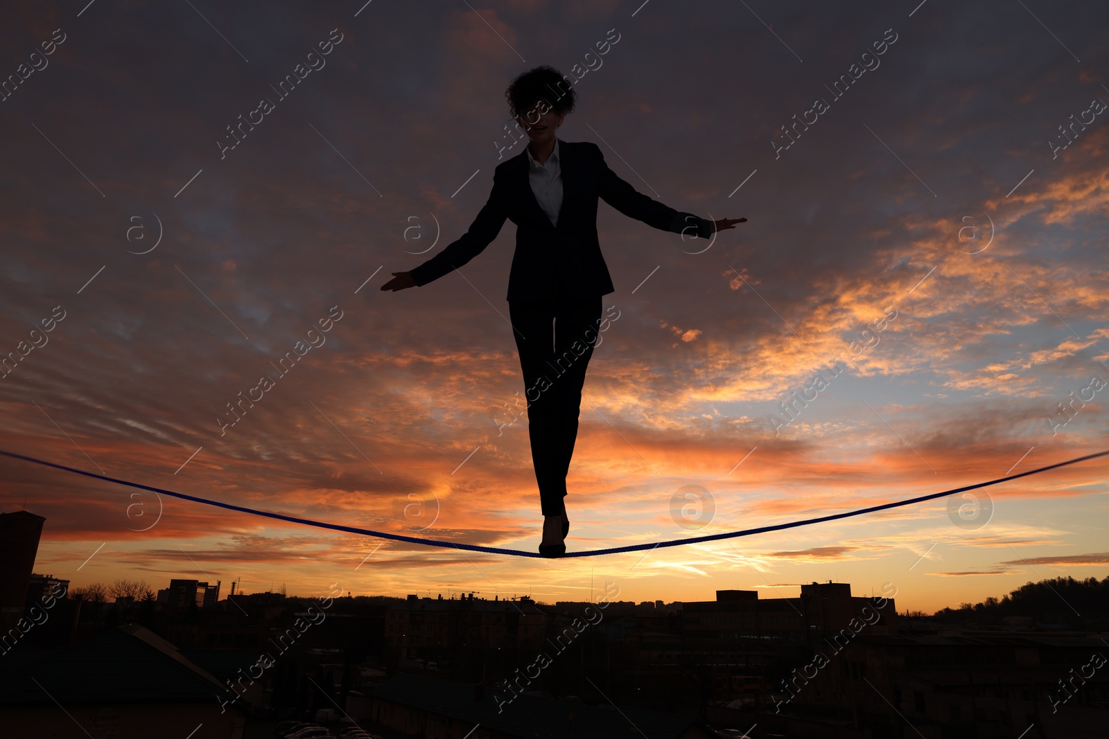 Image of Businesswoman walking rope over city at sunset. Concept of risk and balance