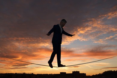 Image of Businessman walking rope over city at sunset. Concept of risk and balance