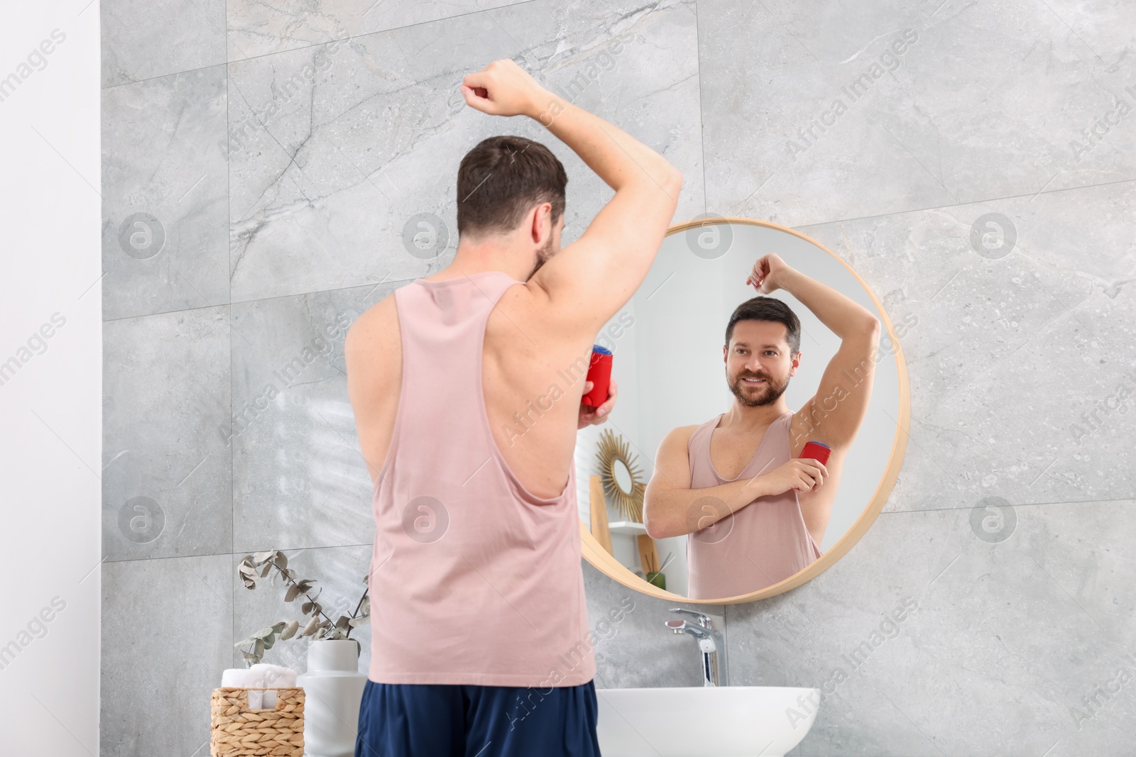 Photo of Smiling man applying solid deodorant near mirror at home, back view