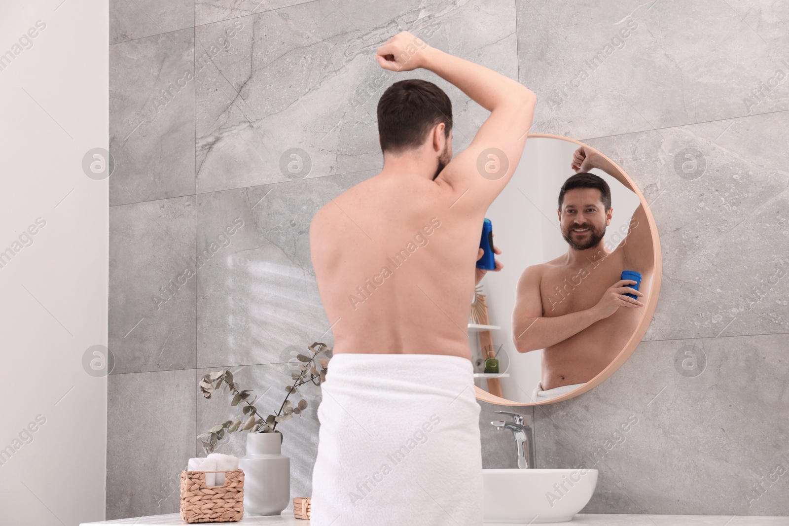 Photo of Smiling man applying solid deodorant near mirror in bathroom, back view