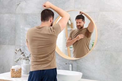 Photo of Emotional man in t-shirt before using deodorant near mirror at home, back view