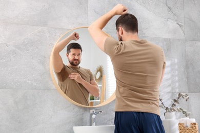 Photo of Emotional man in t-shirt before using deodorant near mirror at home, back view