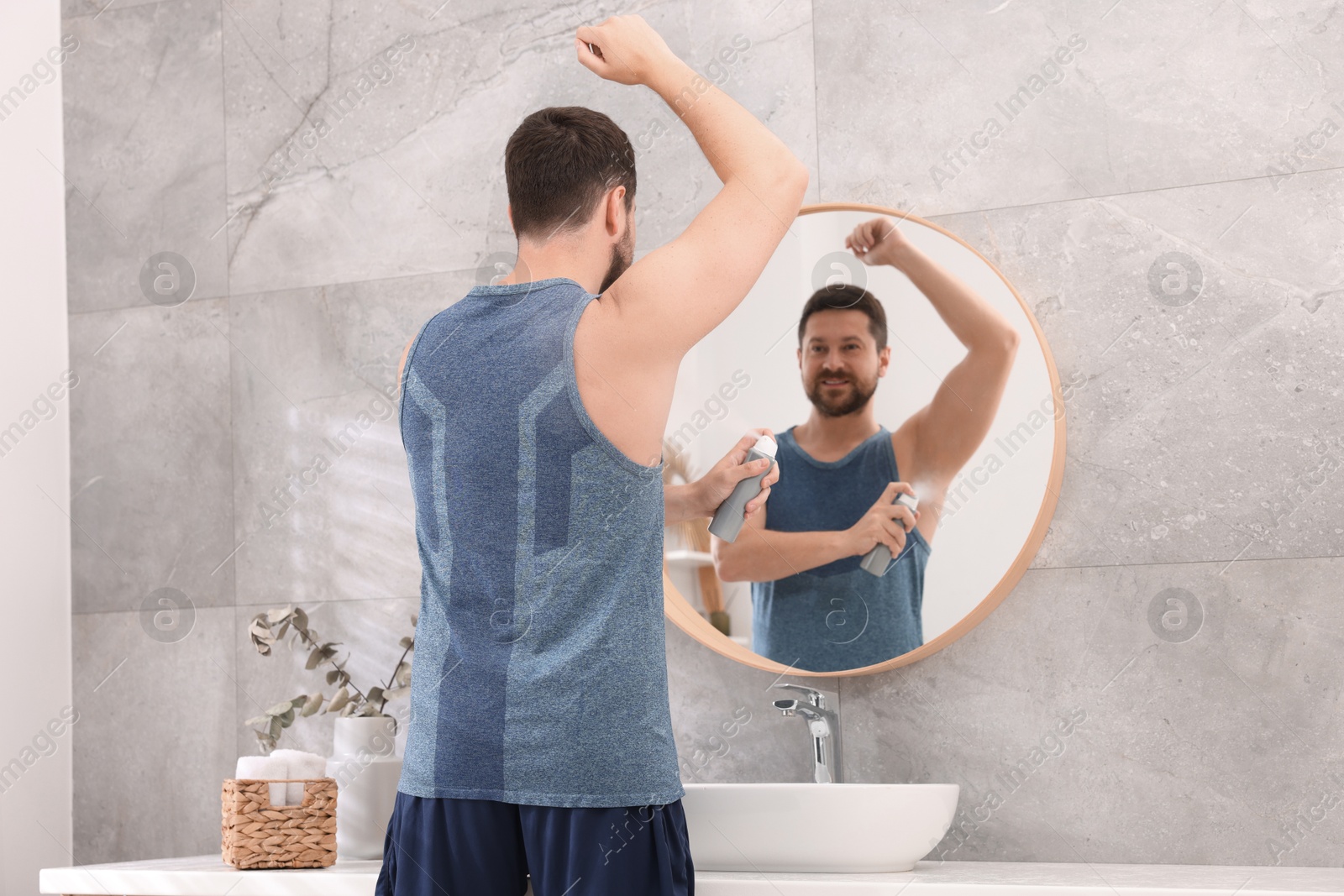 Photo of Smiling man applying spray deodorant near mirror at home, back view