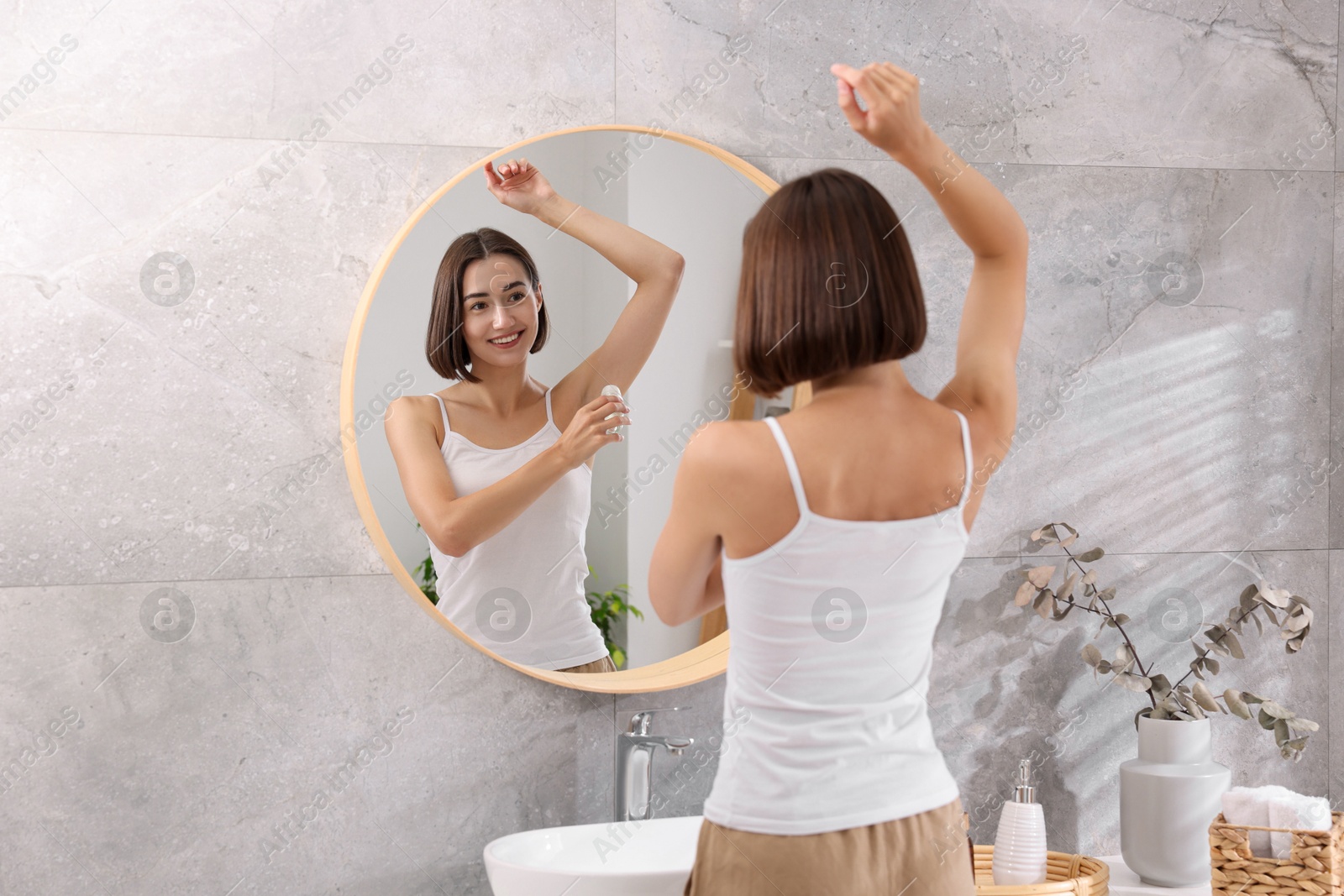 Photo of Smiling woman applying roll-on deodorant near mirror at home, back view