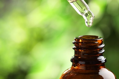 Photo of Essential oil dripping from pipette into bottle against blurred green background, closeup. Space for text