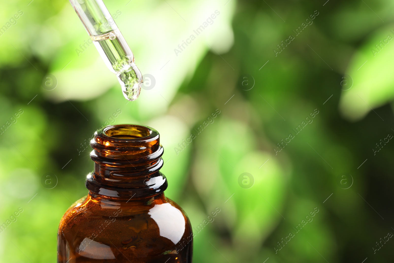 Photo of Essential oil dripping from pipette into bottle against blurred green background, closeup. Space for text