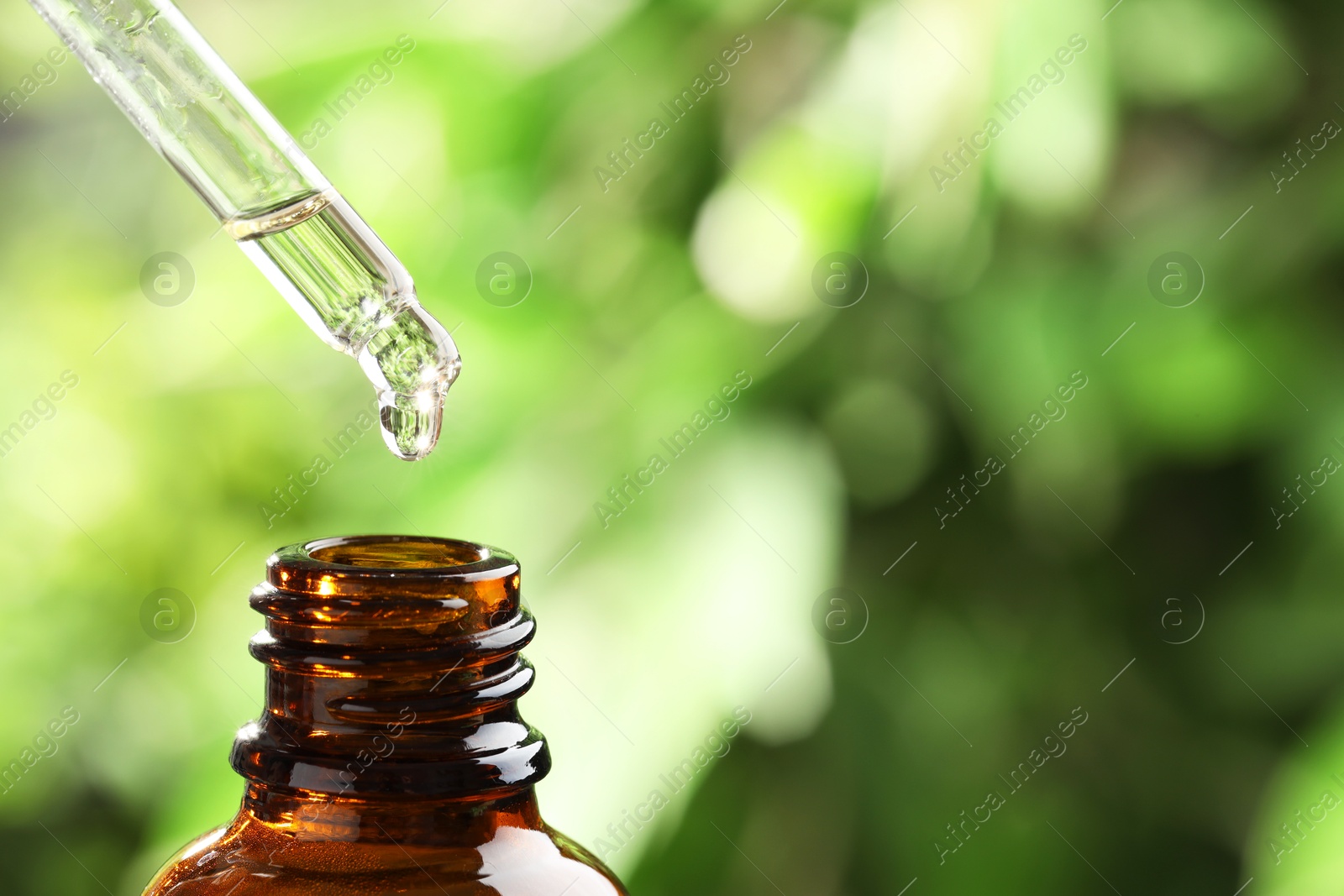 Photo of Essential oil dripping from pipette into bottle against blurred green background, closeup. Space for text