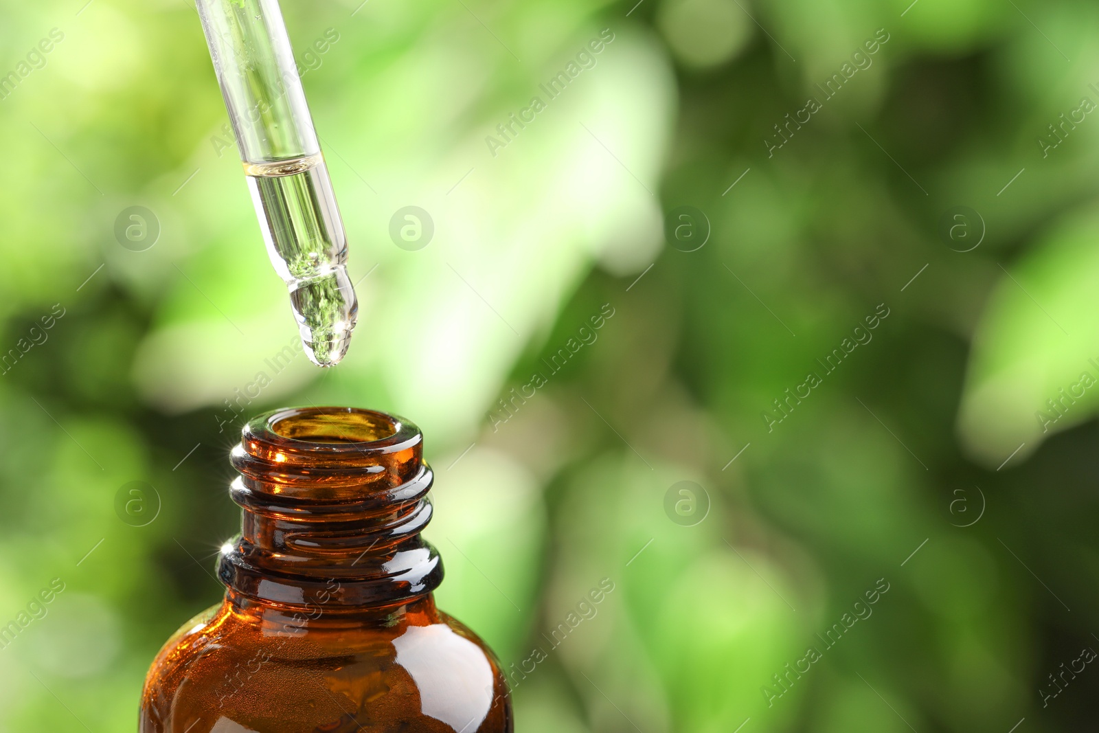 Photo of Essential oil dripping from pipette into bottle against blurred green background, closeup. Space for text