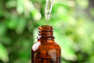 Photo of Essential oil dripping from pipette into bottle against blurred green background, closeup