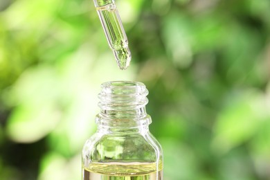 Essential oil dripping from pipette into bottle against blurred green background, closeup. Space for text