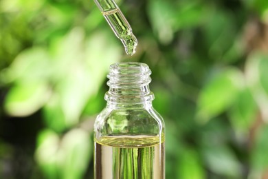 Photo of Essential oil dripping from pipette into bottle against blurred green background, closeup