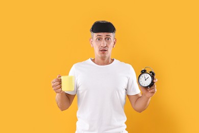 Photo of Overslept man with sleep mask, cup and alarm clock on orange background