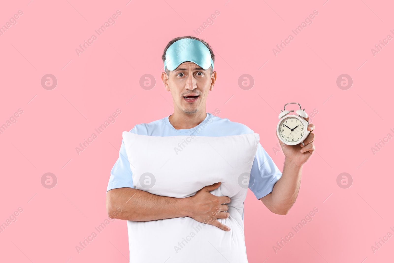 Photo of Overslept man with sleep mask, pillow and alarm clock on pink background