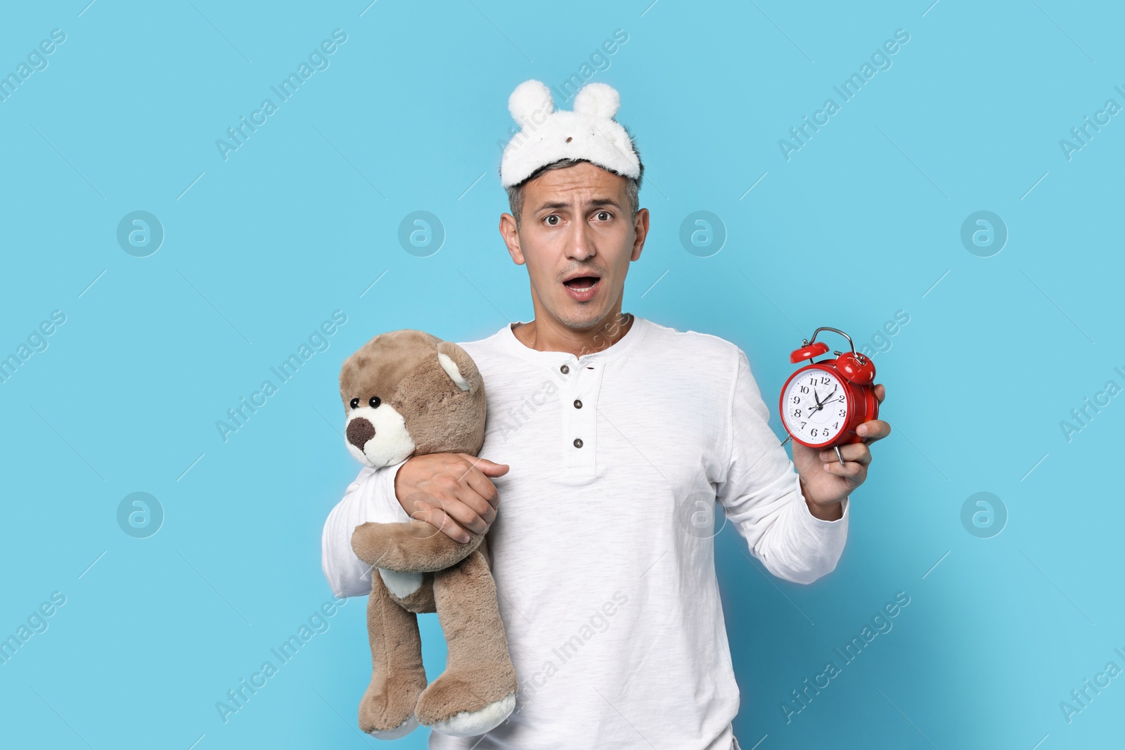 Photo of Overslept man with sleep mask, teddy bear and alarm clock on light blue background