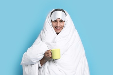 Happy man with sleep mask and cup wrapped in blanket on light blue background