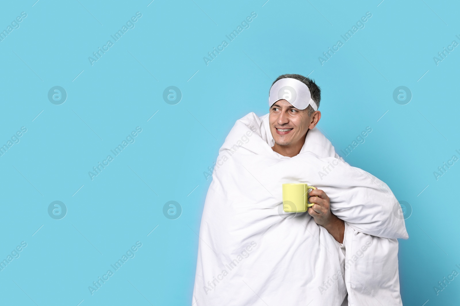 Photo of Happy man with sleep mask and cup wrapped in blanket on light blue background, space for text