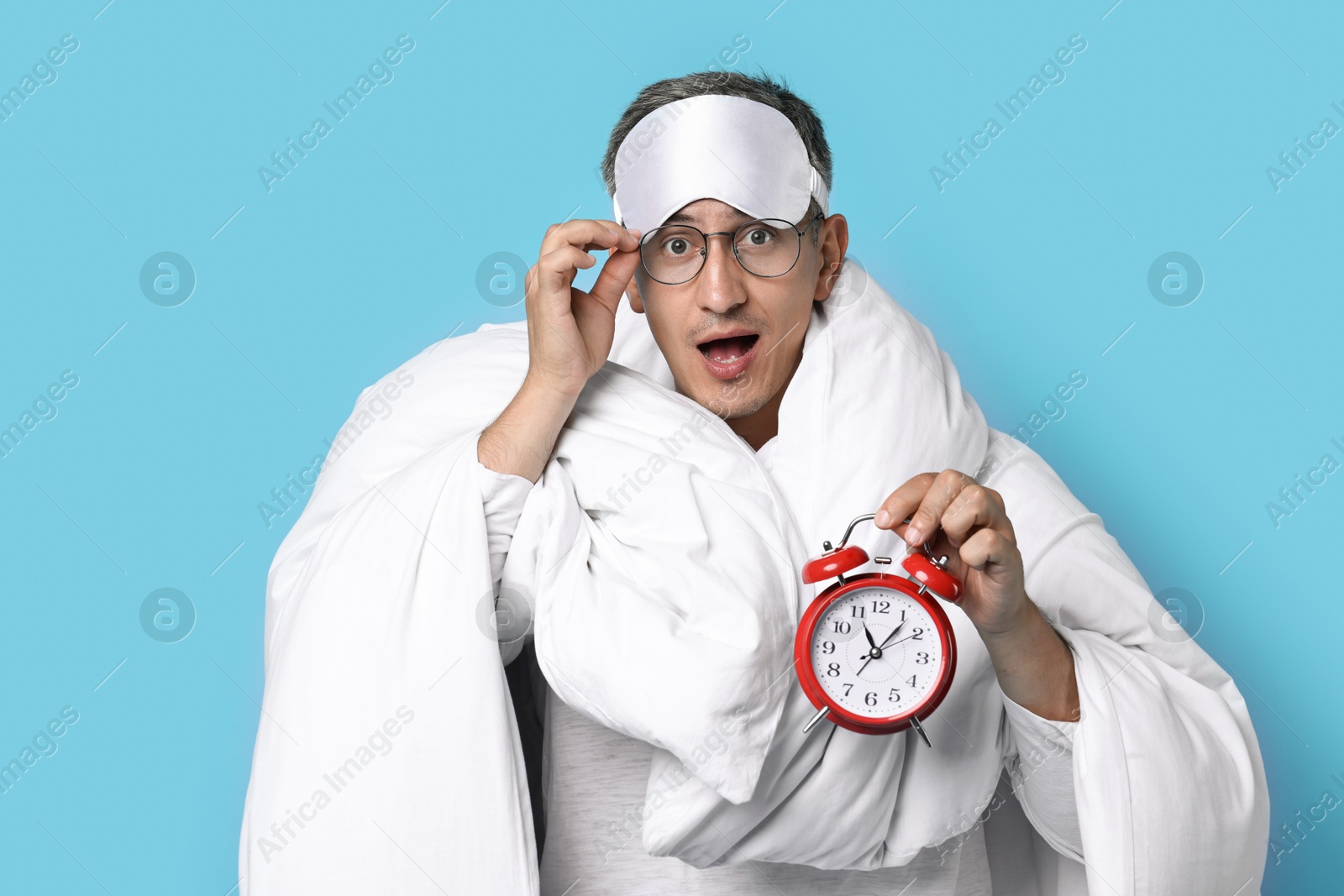 Photo of Overslept man with sleep mask and alarm clock wrapped in blanket on light blue background