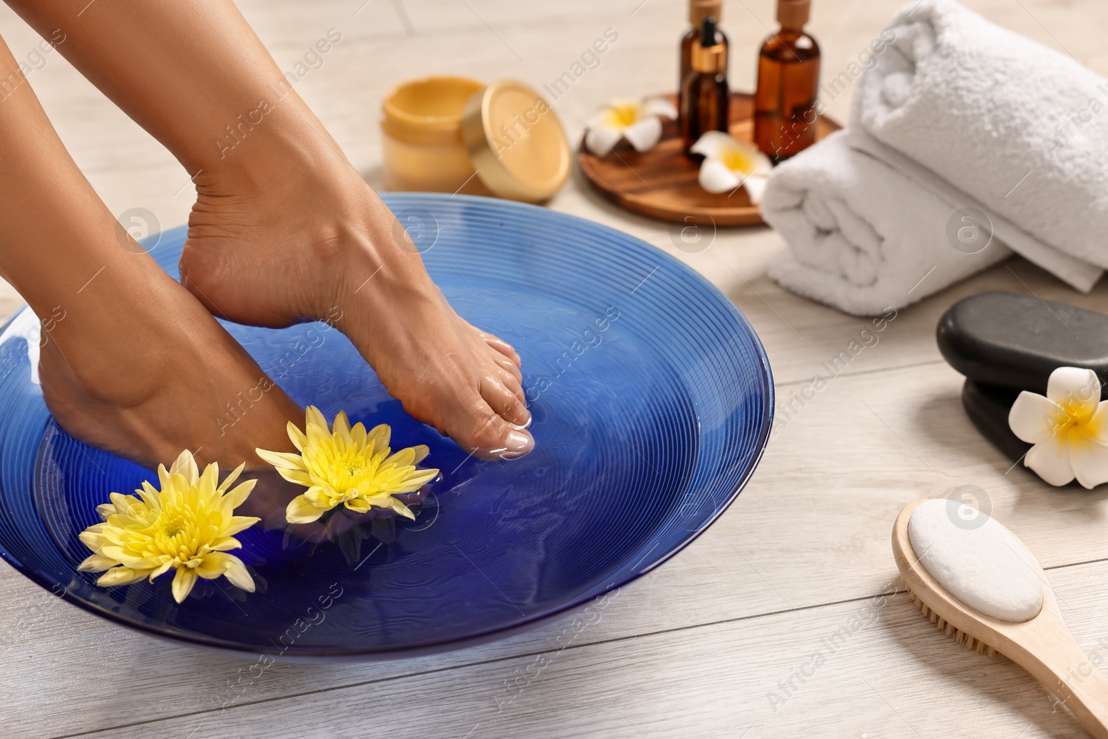 Photo of Woman soaking her feet in bowl with water and flowers on floor, closeup. Body care