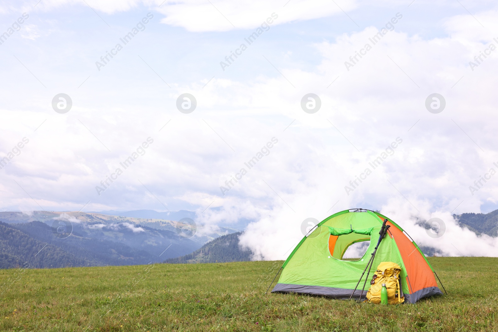 Photo of Tent, backpack, trekking poles and thermos on green grass in mountains, space for text