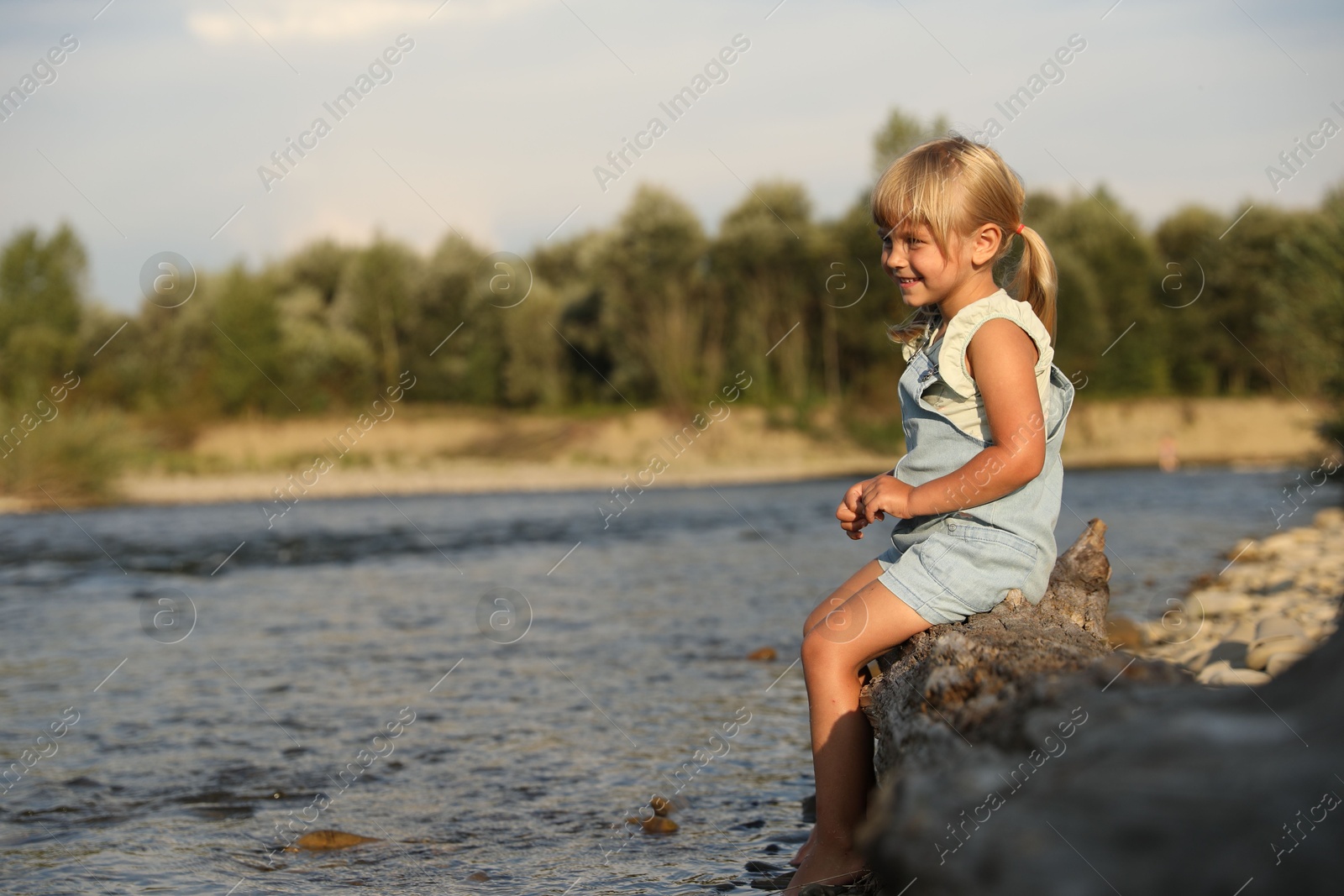 Photo of Cute little girl sitting on tree trunk near river. Child enjoying beautiful nature