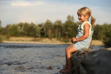 Cute little girl sitting on tree trunk near river. Child enjoying beautiful nature