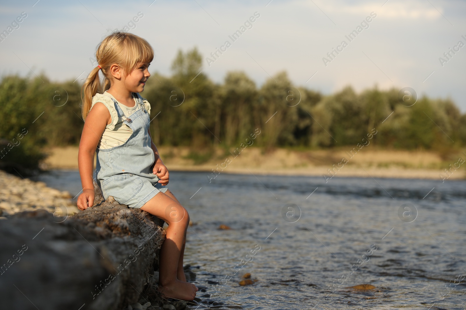 Photo of Cute little girl sitting on tree trunk near river. Child enjoying beautiful nature