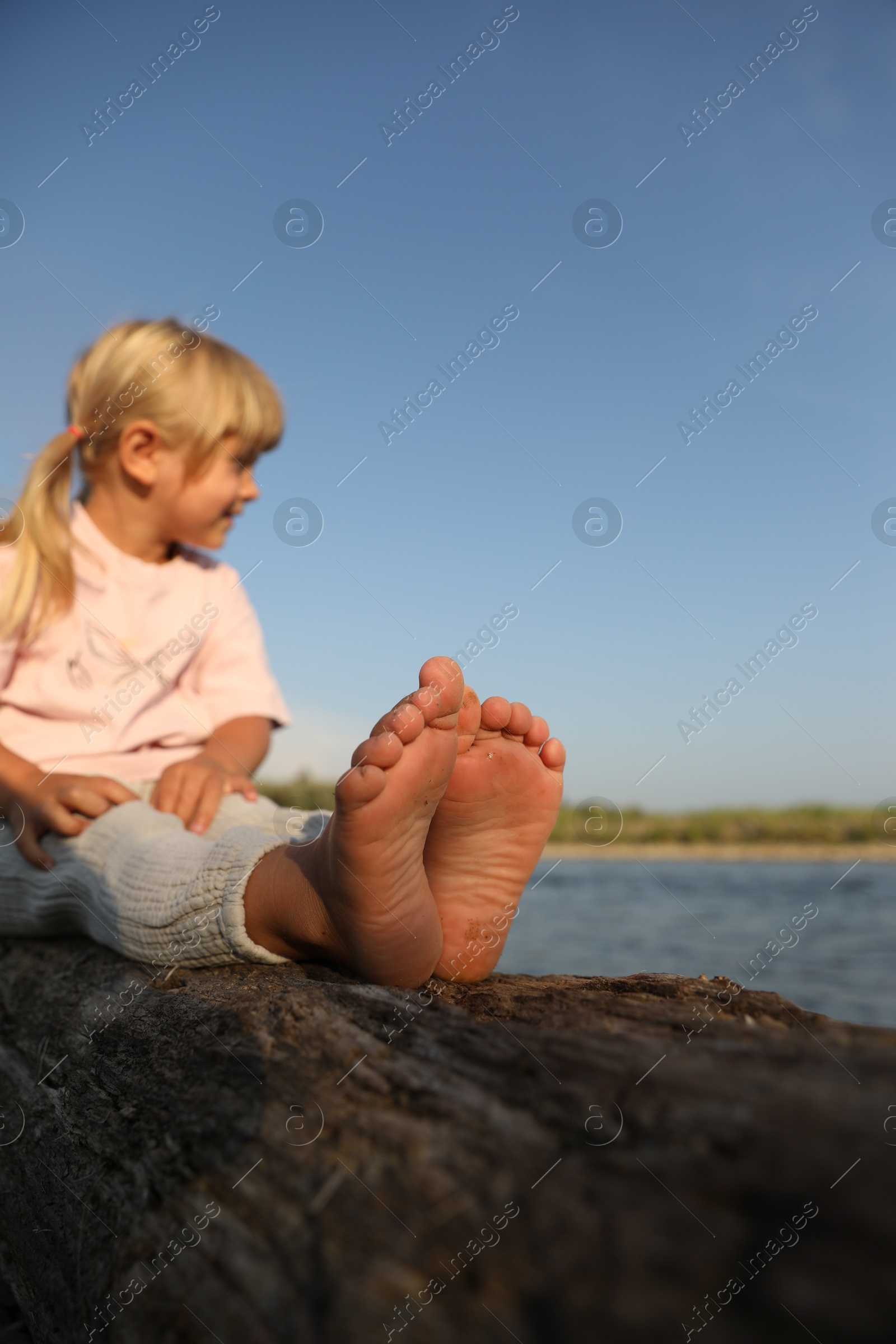 Photo of Cute little girl sitting on tree trunk near river, selective focus. Child enjoying beautiful nature