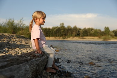 Cute little girl sitting on tree trunk near river. Child enjoying beautiful nature