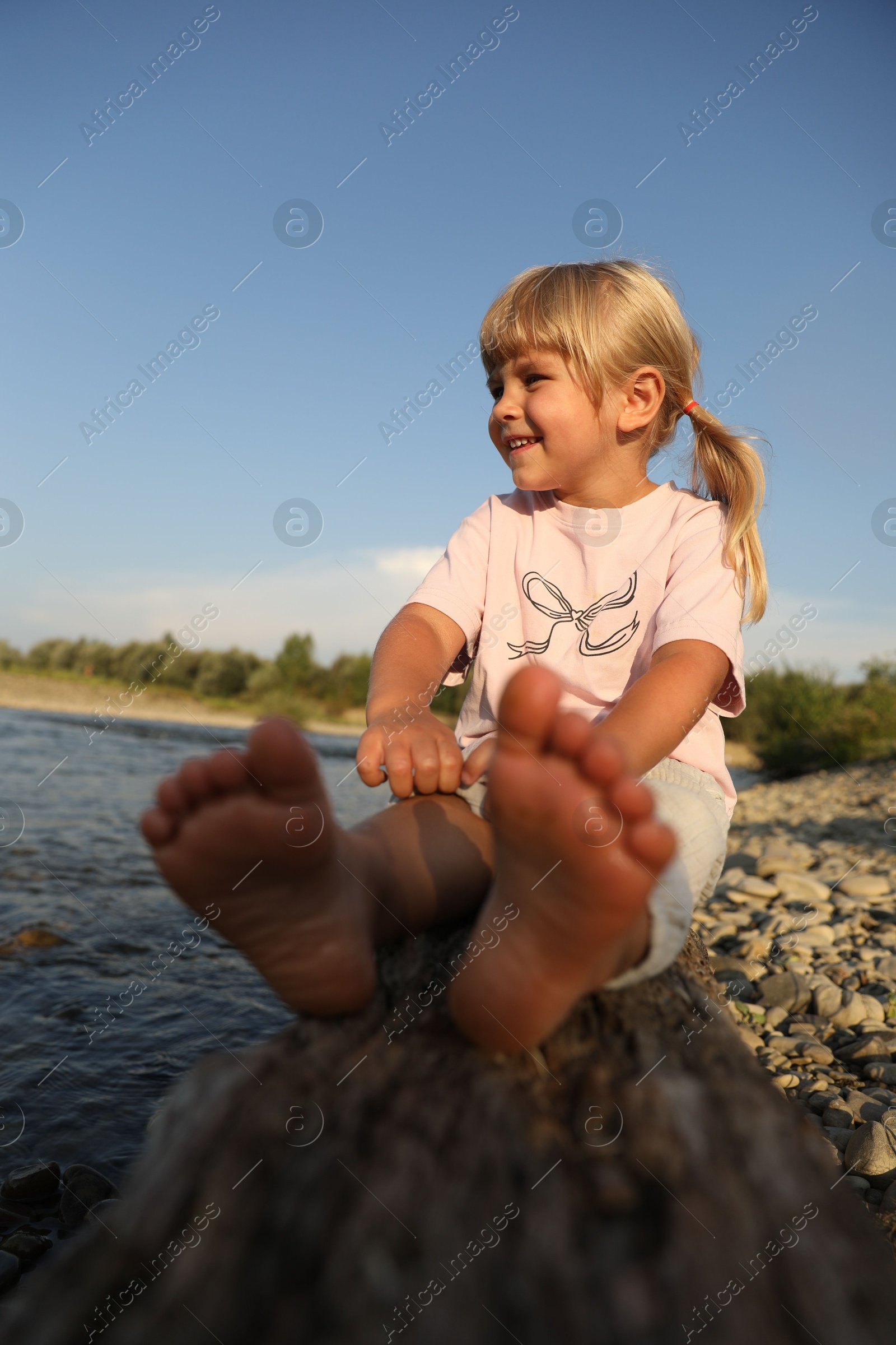 Photo of Cute little girl sitting on tree trunk near river. Child enjoying beautiful nature