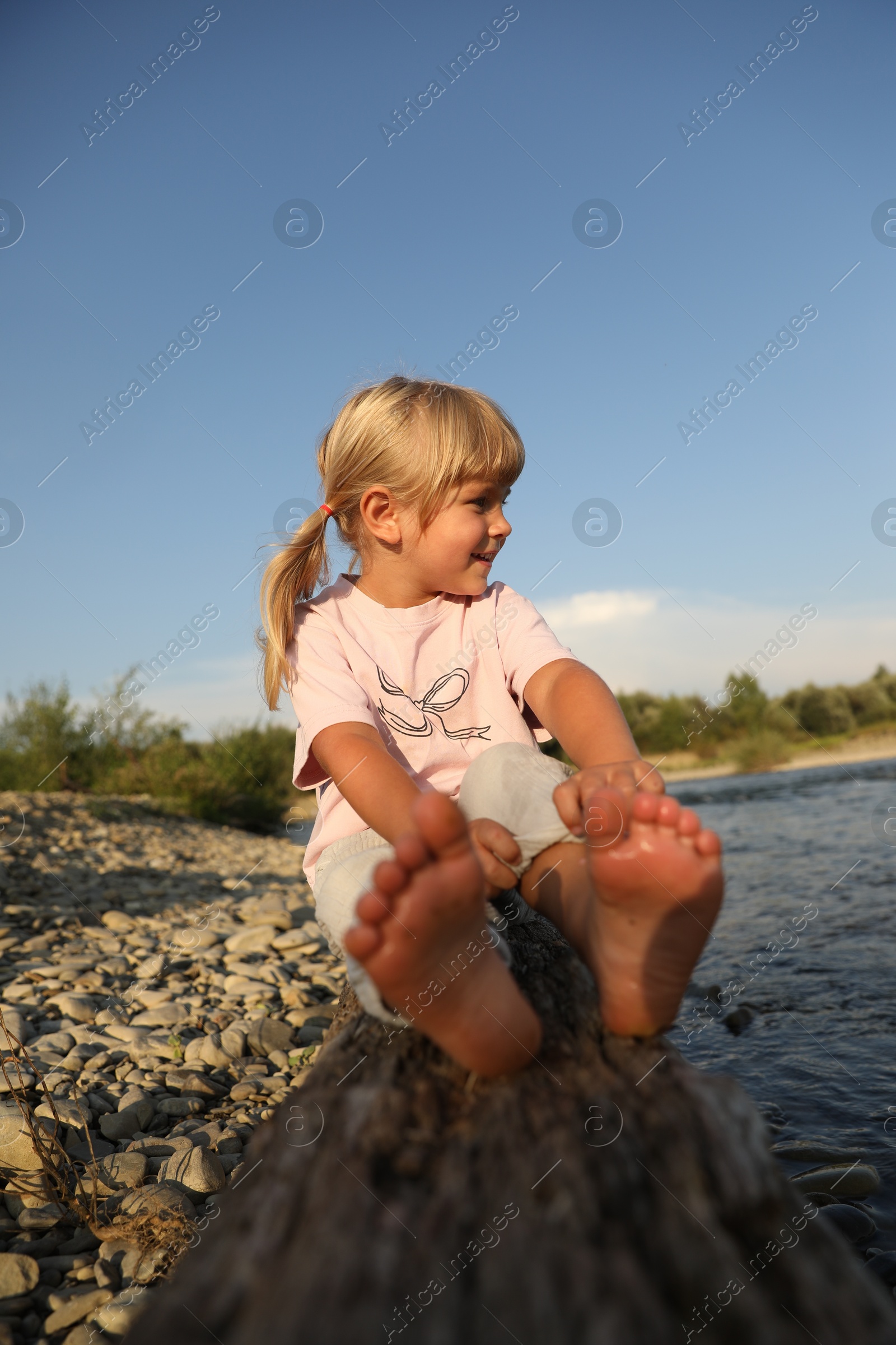 Photo of Cute little girl sitting on tree trunk near river. Child enjoying beautiful nature