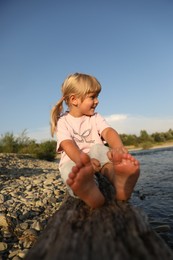 Photo of Cute little girl sitting on tree trunk near river. Child enjoying beautiful nature