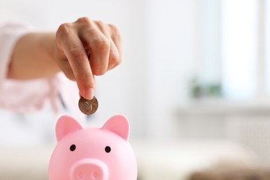 Photo of Woman putting coin into pink piggy bank at home, closeup. Space for text