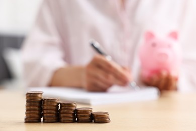 Photo of Woman with piggy bank taking notes at table indoors, focus on coins