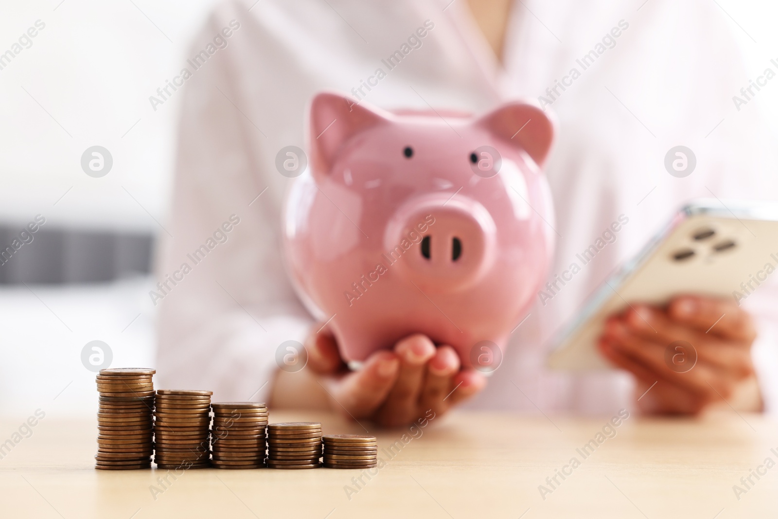 Photo of Woman with piggy bank and smartphone at table indoors, focus on coins