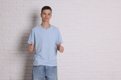 Photo of Teenage boy wearing light blue t-shirt and showing thumbs up near white brick wall, space for text