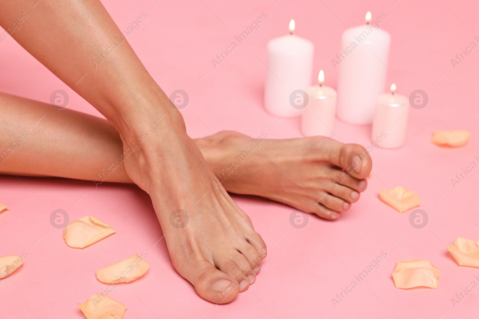 Photo of Woman with smooth feet, burning candles and petals on pink background, closeup