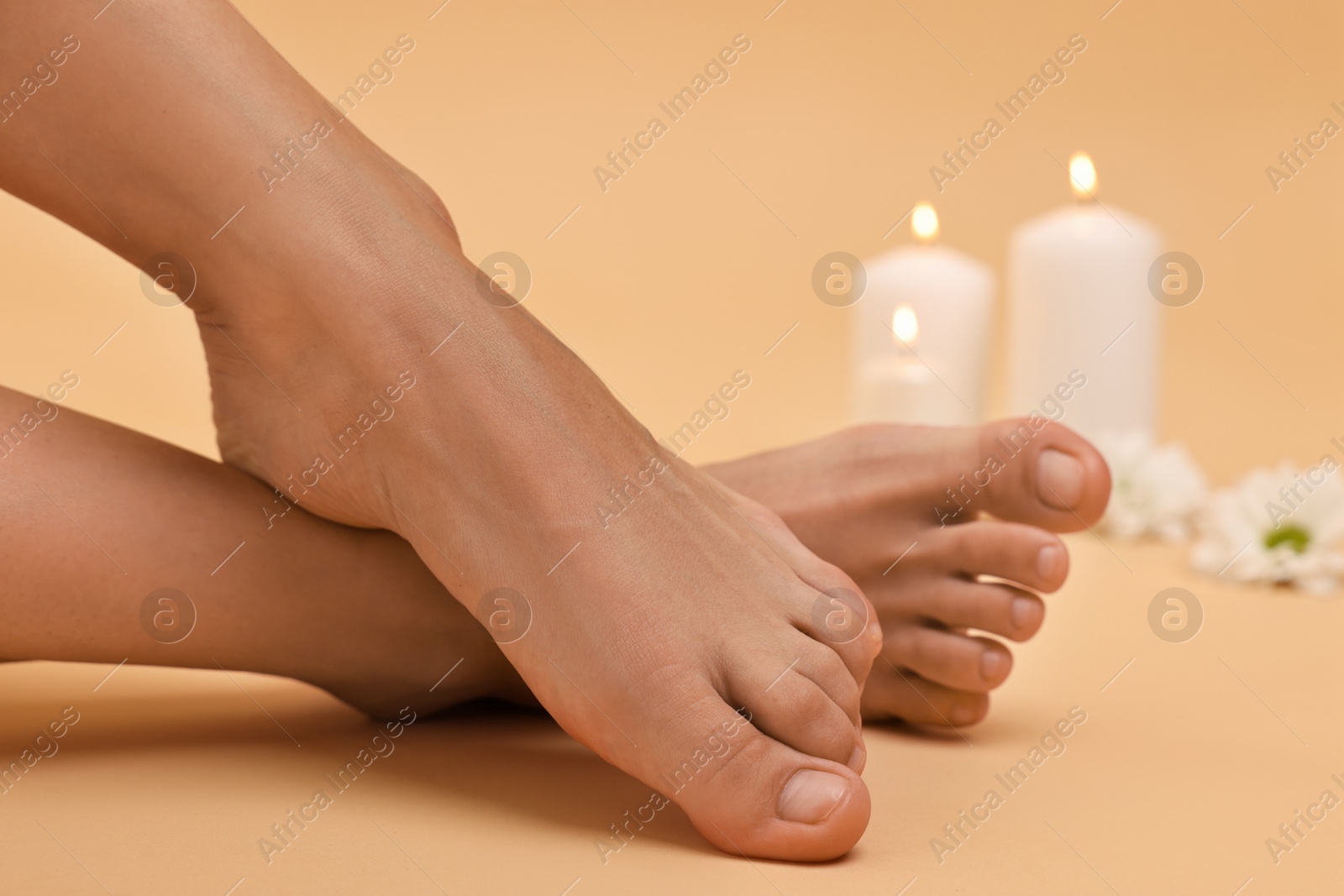 Photo of Woman with smooth feet, burning candles and chrysanthemum flowers on beige background, closeup