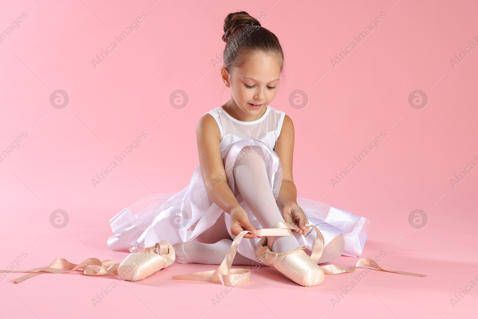 Photo of Little ballerina tying pointe shoes on pink background