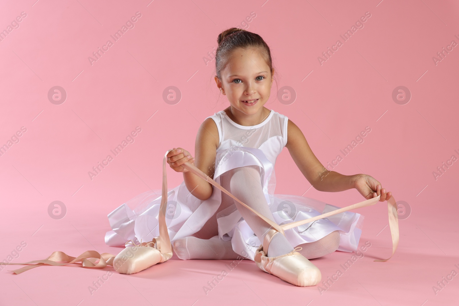 Photo of Little ballerina tying pointe shoes on pink background