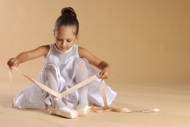 Little ballerina tying pointe shoes on beige background, space for text