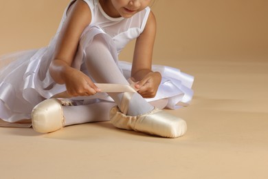 Photo of Little ballerina tying pointe shoes on beige background, closeup