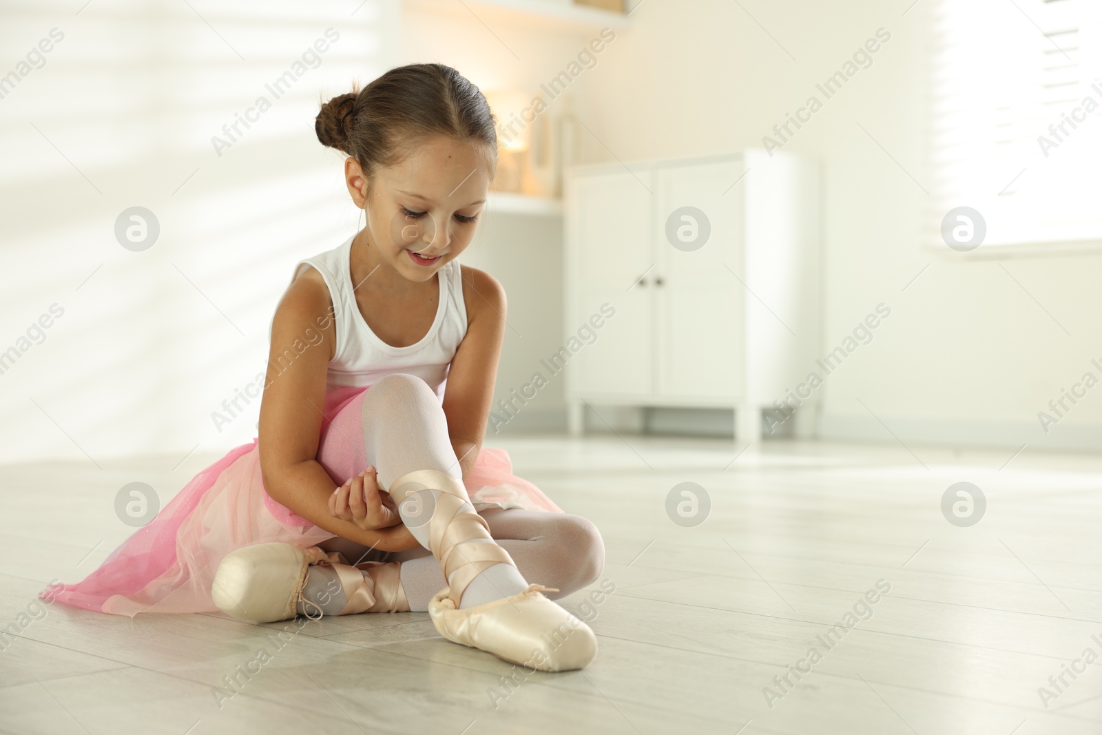 Photo of Little ballerina tying pointe shoes indoors, space for text