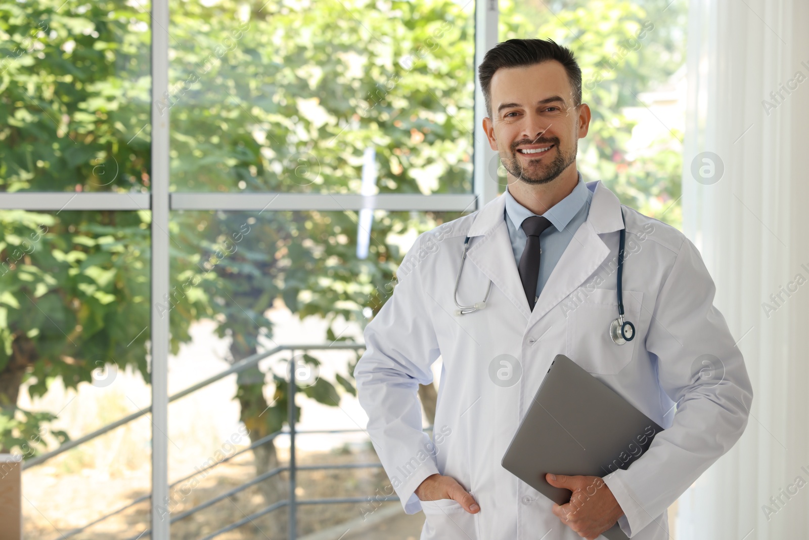 Photo of Smiling doctor with stethoscope and laptop indoors, space for text