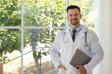 Smiling doctor with stethoscope and laptop indoors, space for text