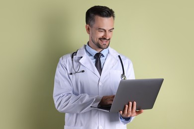 Smiling doctor with stethoscope using laptop on olive background