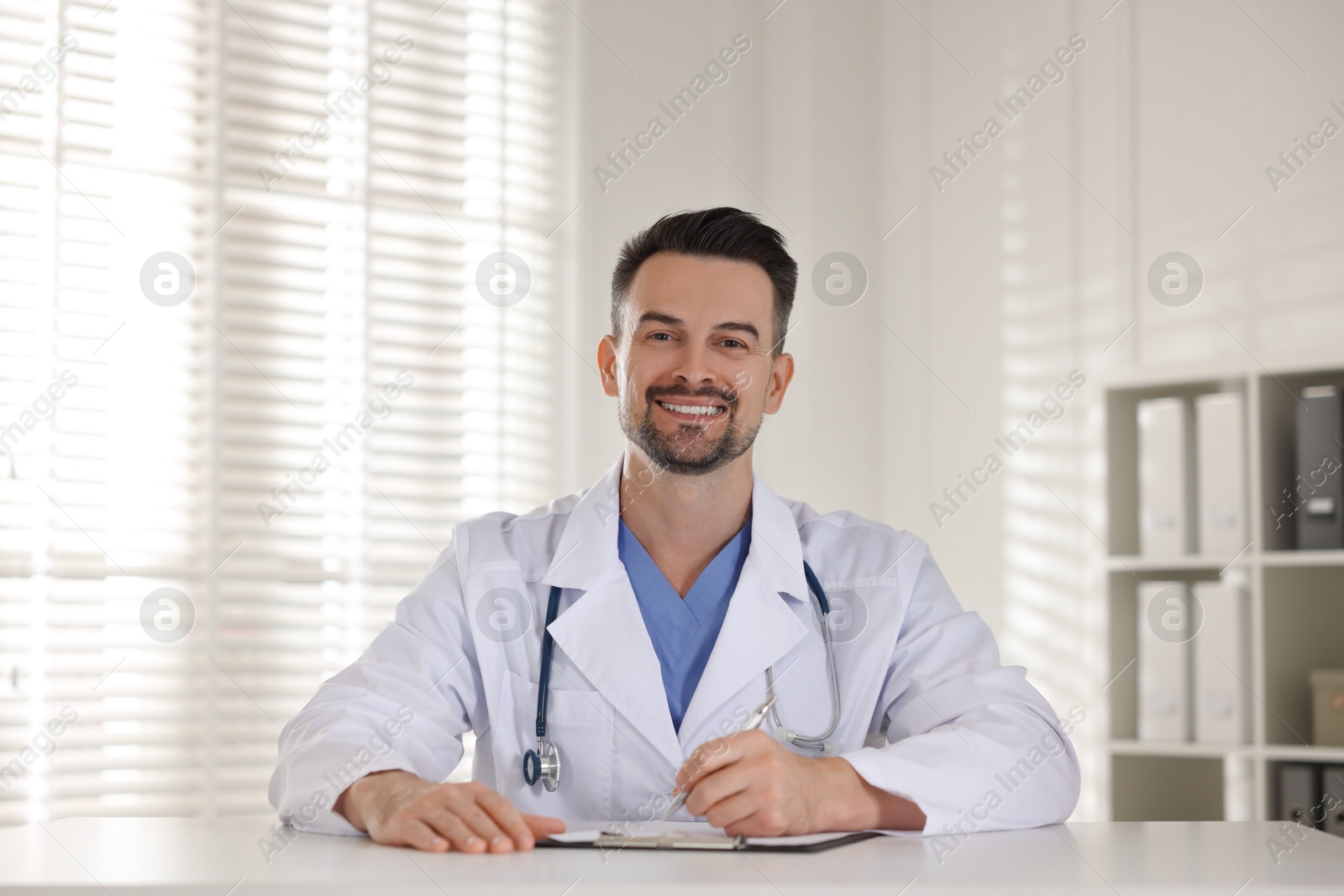 Photo of Smiling doctor with clipboard at table in clinic