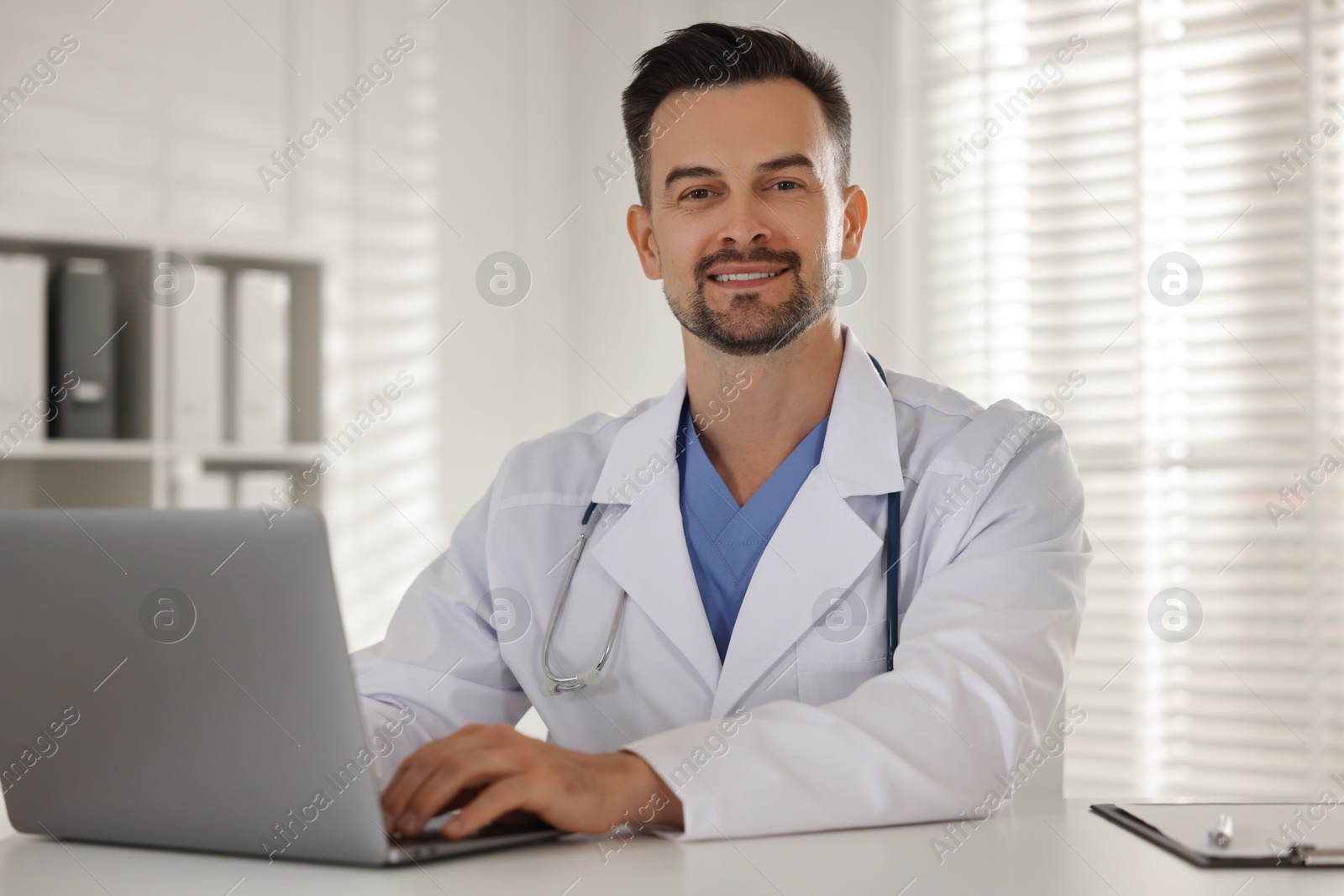 Photo of Smiling doctor working with laptop at table in clinic